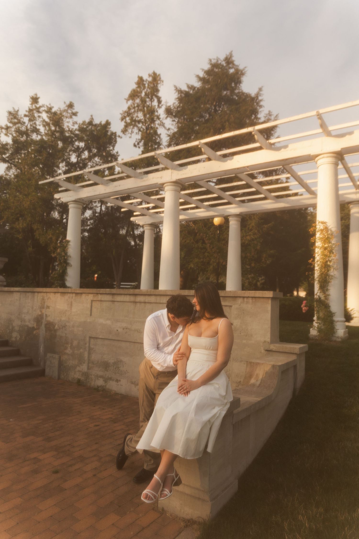Couple in white attire sitting intimately on stone bench under elegant white pergola at golden hour in lush garden.