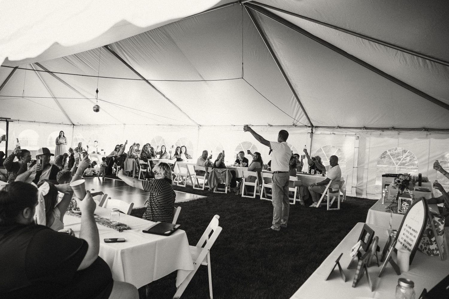 Black and white photo of guests raising glasses during a toast at an elegant outdoor wedding reception under a large tent.