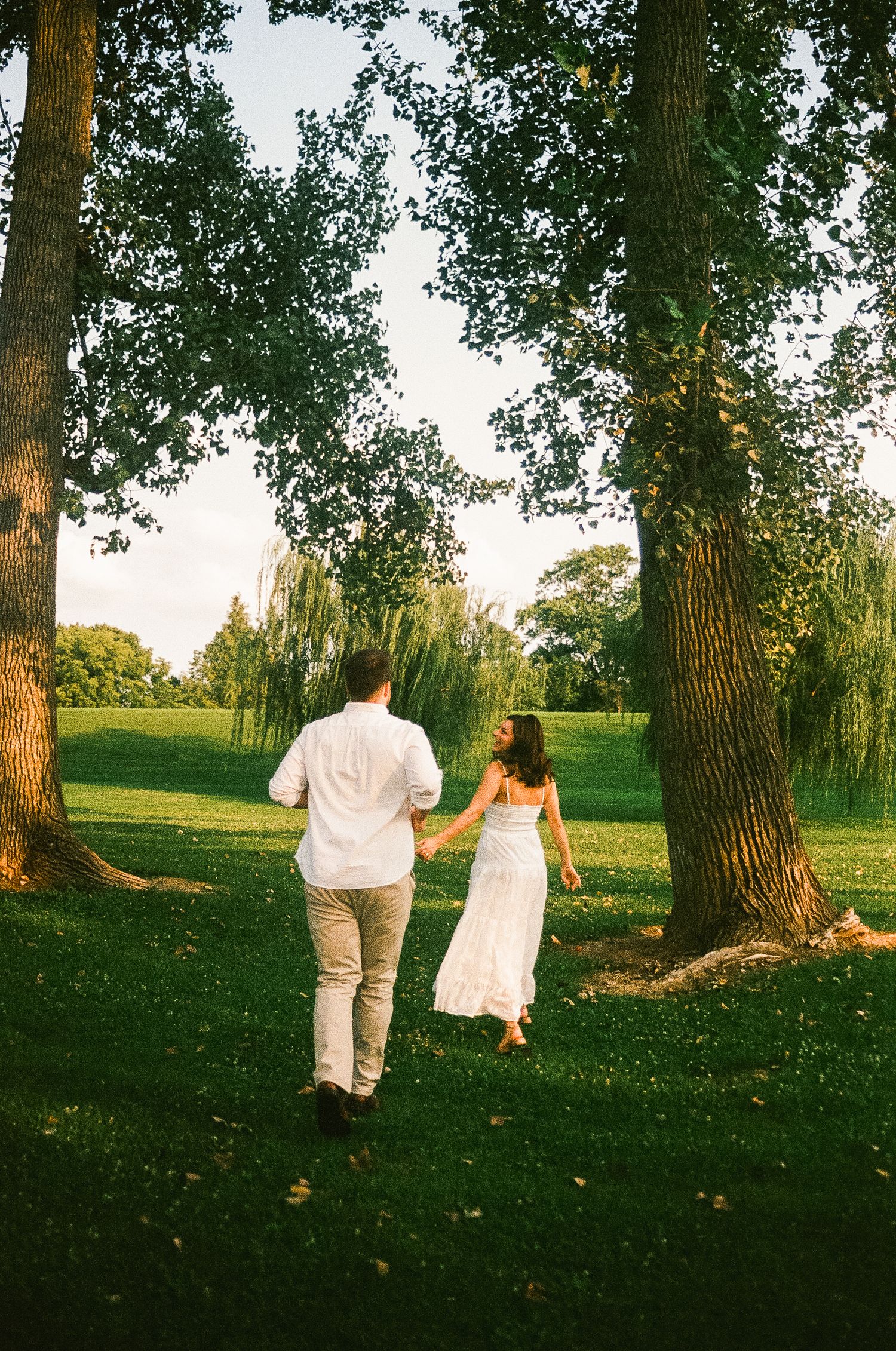 Couple holding hands walking through lush green park with tall trees during golden hour.