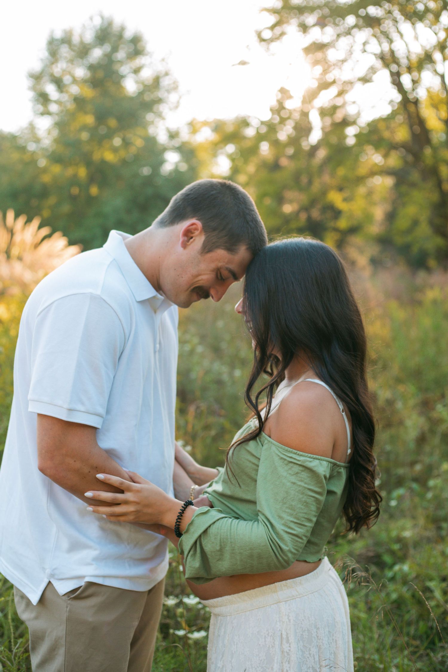 Tender maternity moment as couple bows heads together, hands cradling pregnant belly in sunlit field.