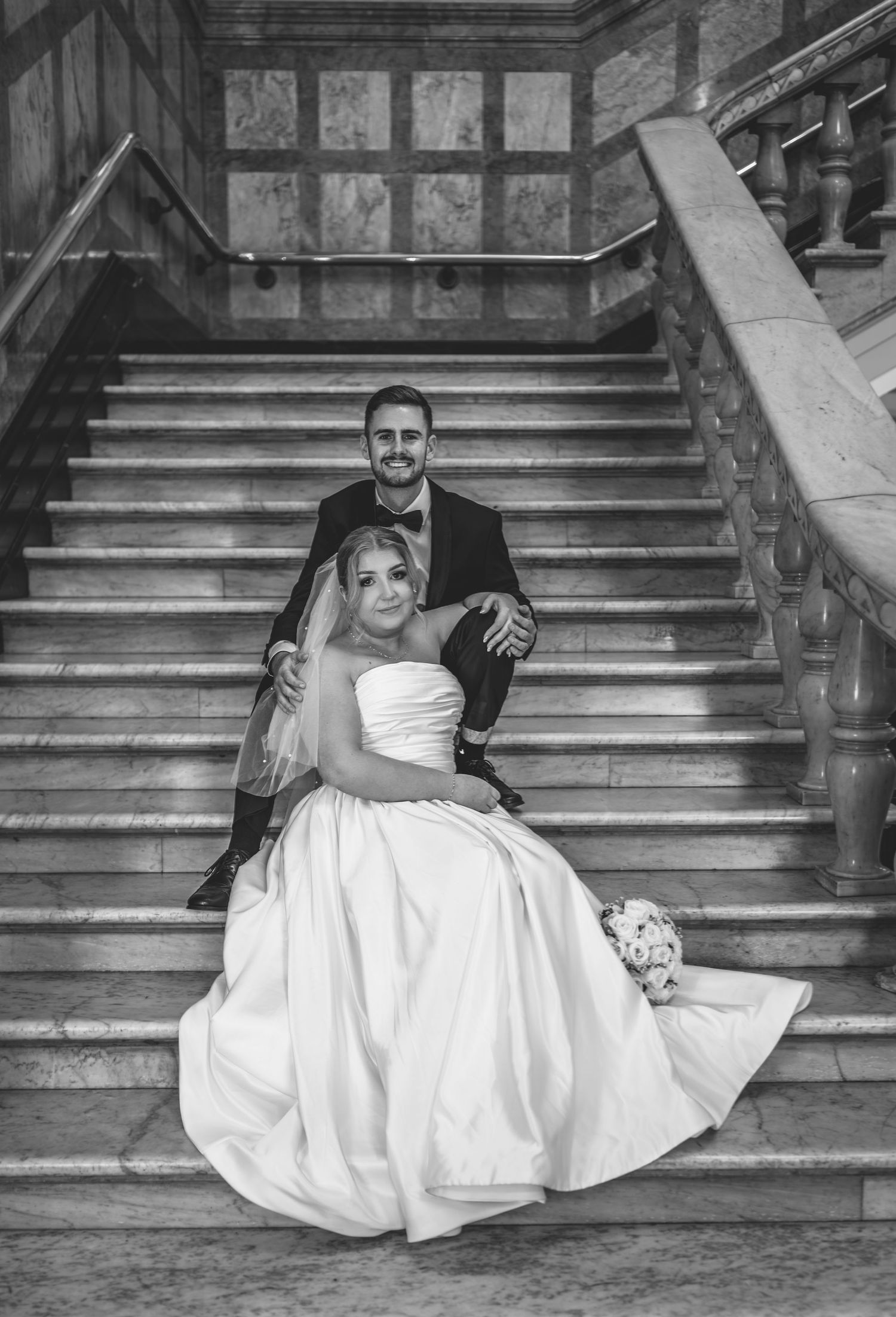 A black and white wedding photograph showing a couple seated on elegant stone stairs with the bride in a white gown.