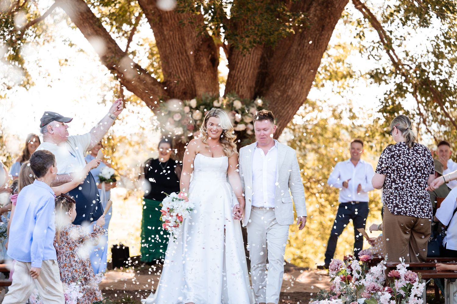 Bride and groom walk through confetti shower under large oak tree surrounded by smiling wedding guests outdoors.
