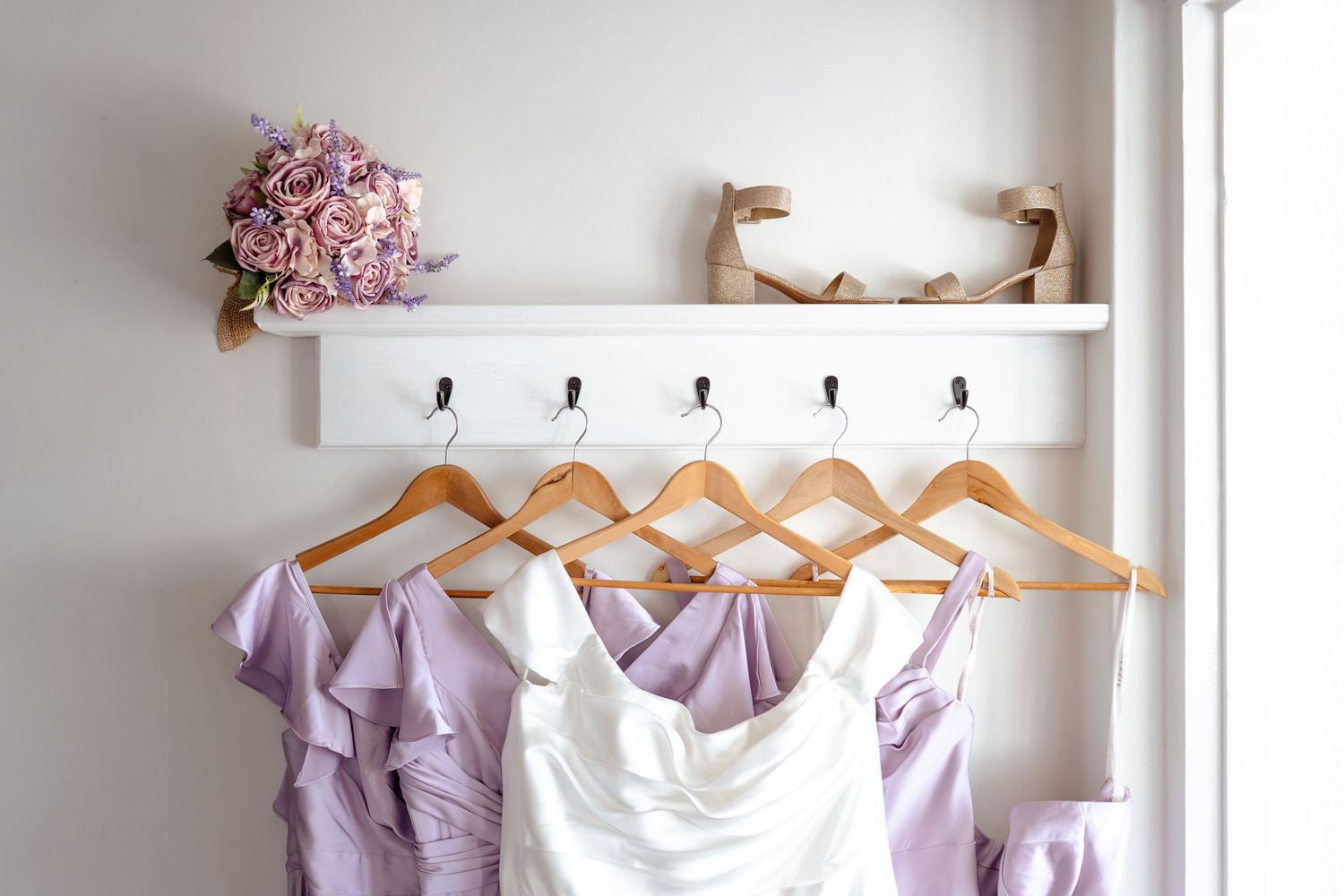 White wedding dress and lavender bridesmaid dresses hanging on wooden hangers with floral bouquet on shelf.