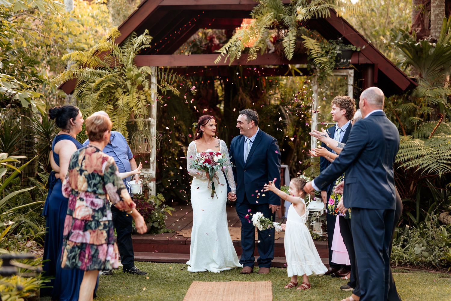Bride and groom walk through confetti shower at outdoor garden wedding ceremony with guests celebrating.