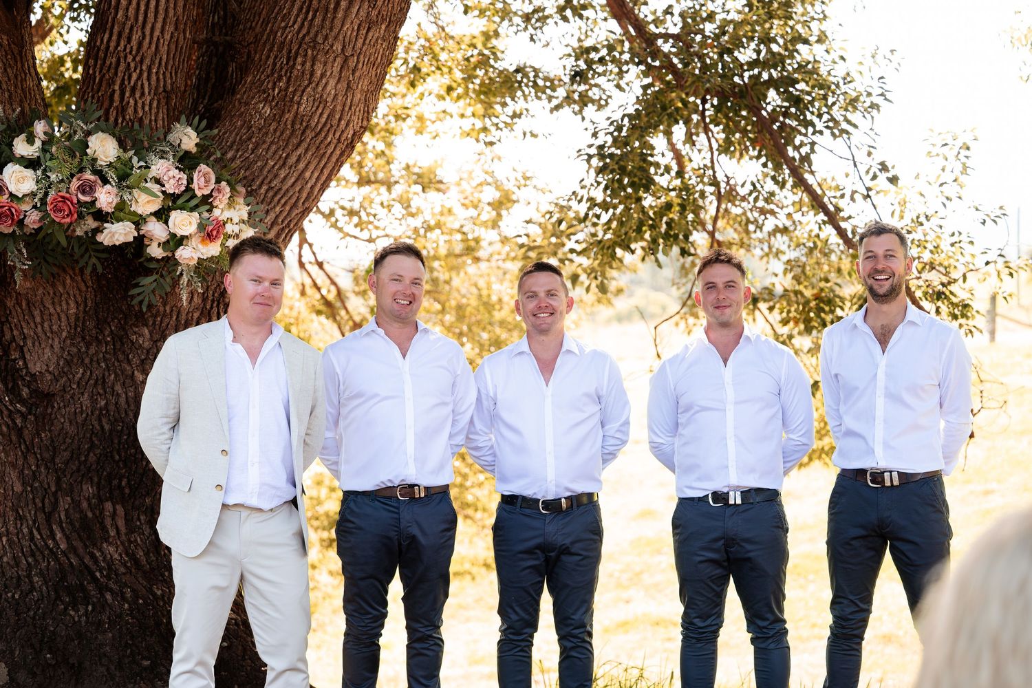 Five groomsmen in white shirts and navy pants smiling outdoors near a floral arrangement on a tree.