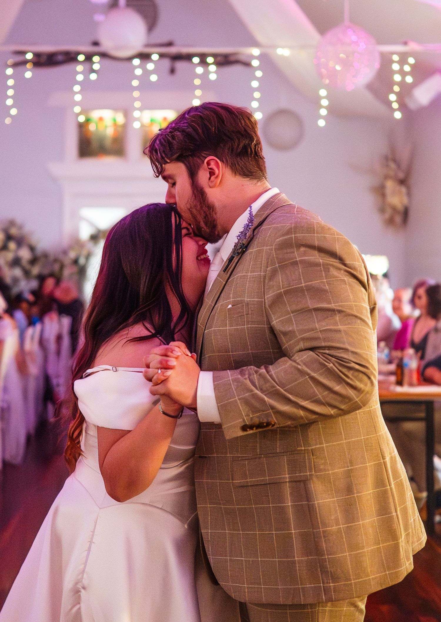 Bride and groom share first dance kiss at wedding reception, man in plaid suit, woman in white gown.