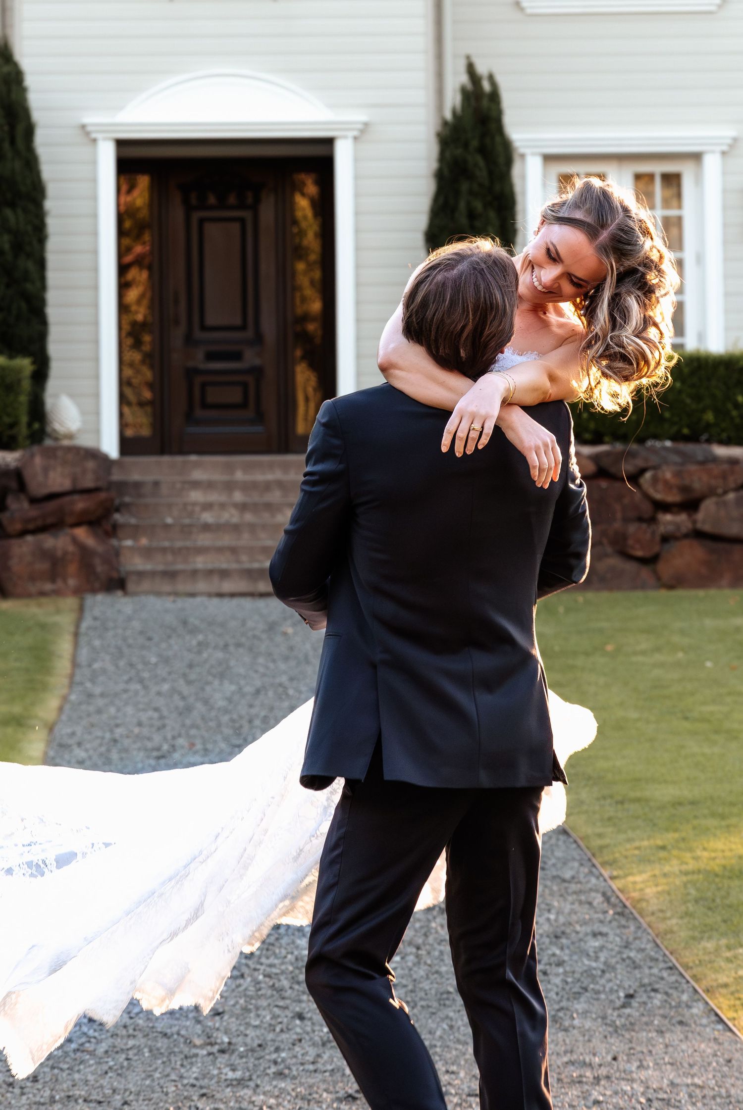 Groom lifting bride in white gown outside elegant white house during golden hour wedding portrait.