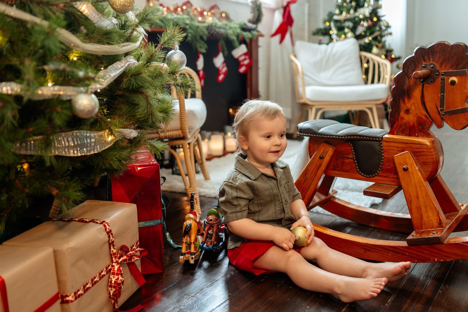 Toddler sitting near a decorated Christmas tree with gifts and a wooden rocking horse on a hardwood floor.