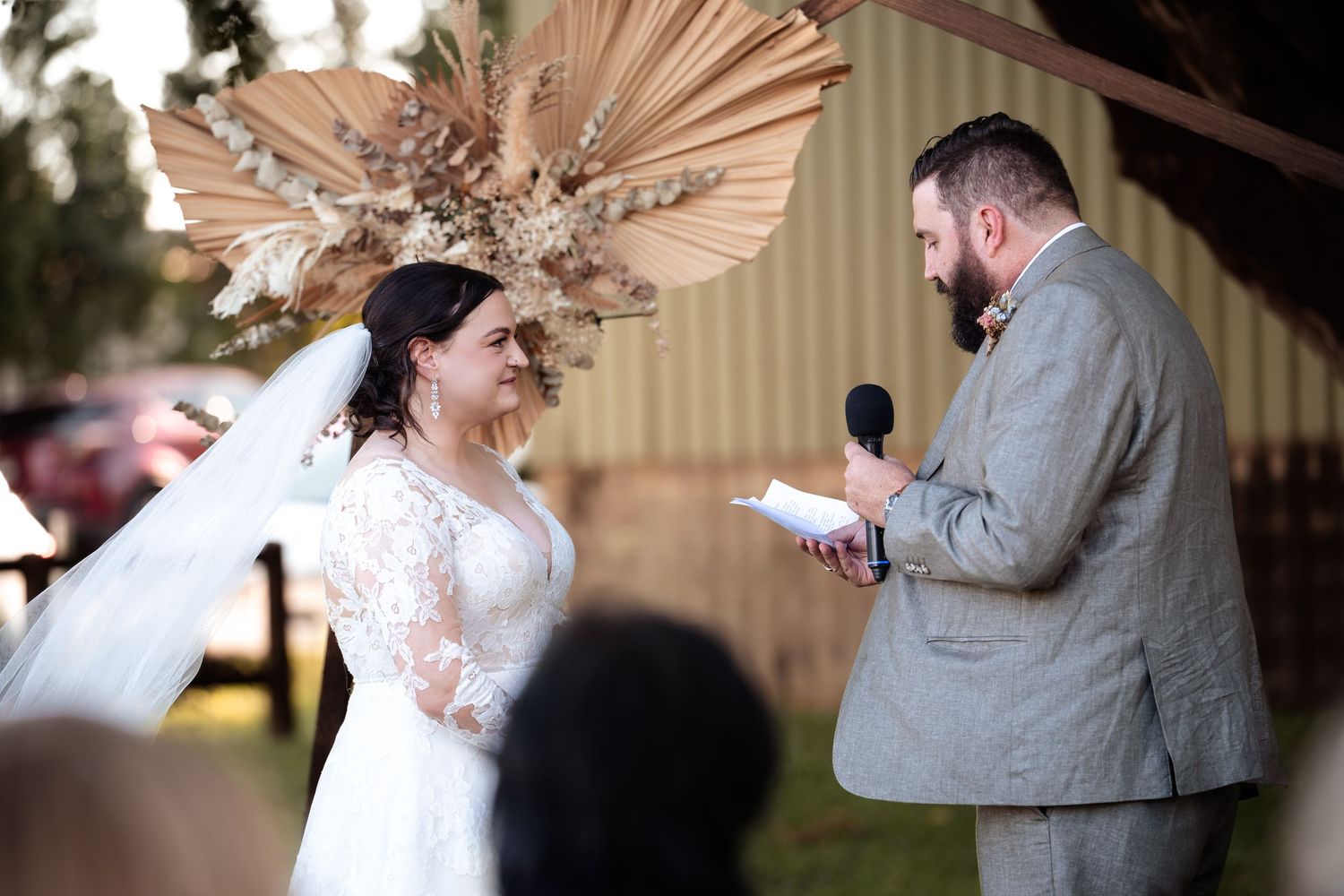 Bride in lace gown and floral headpiece listens as groom reads vows at outdoor rustic wedding ceremony.
