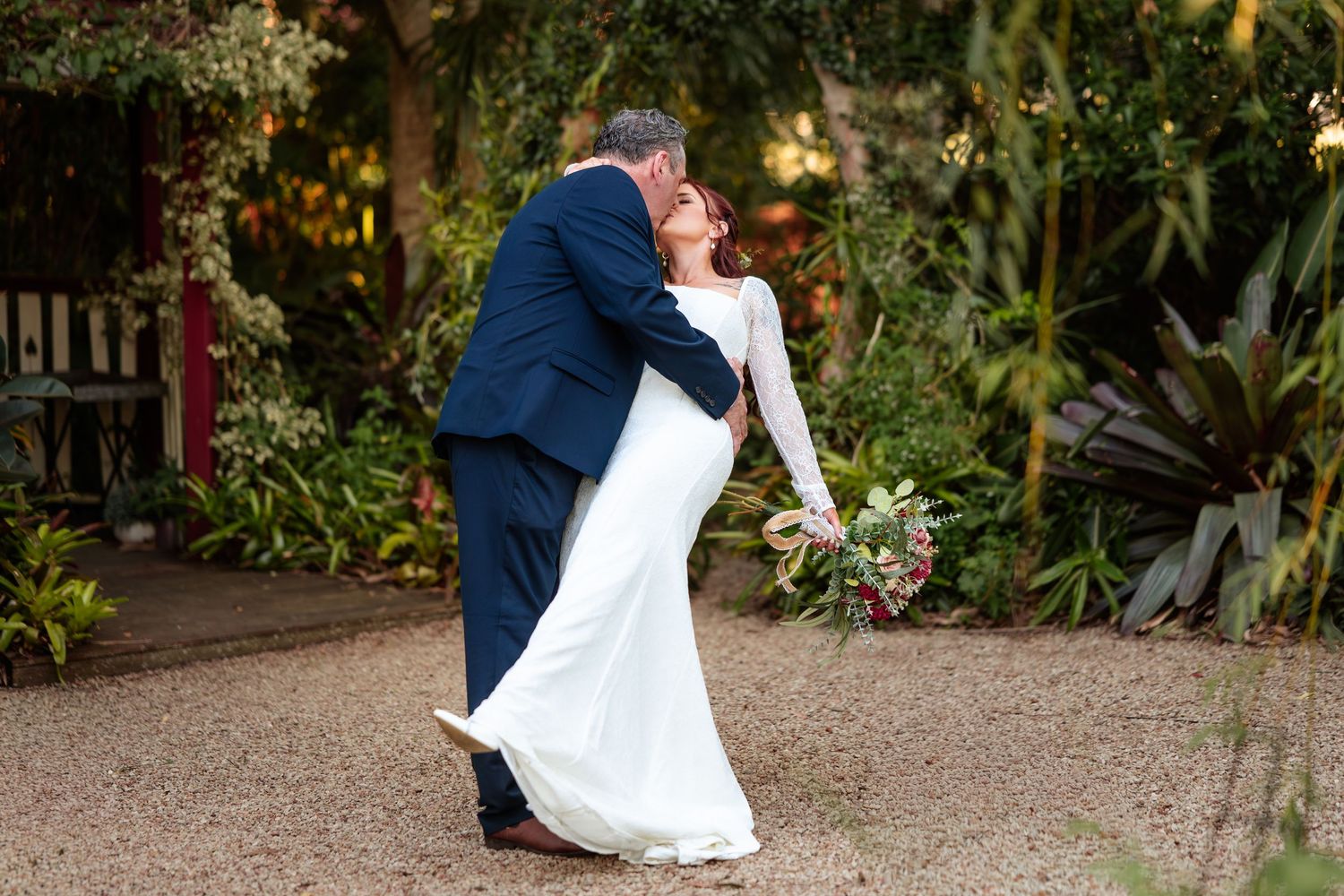 Bride and groom share a romantic dip kiss in a lush garden setting, bride in white gown holding bouquet.