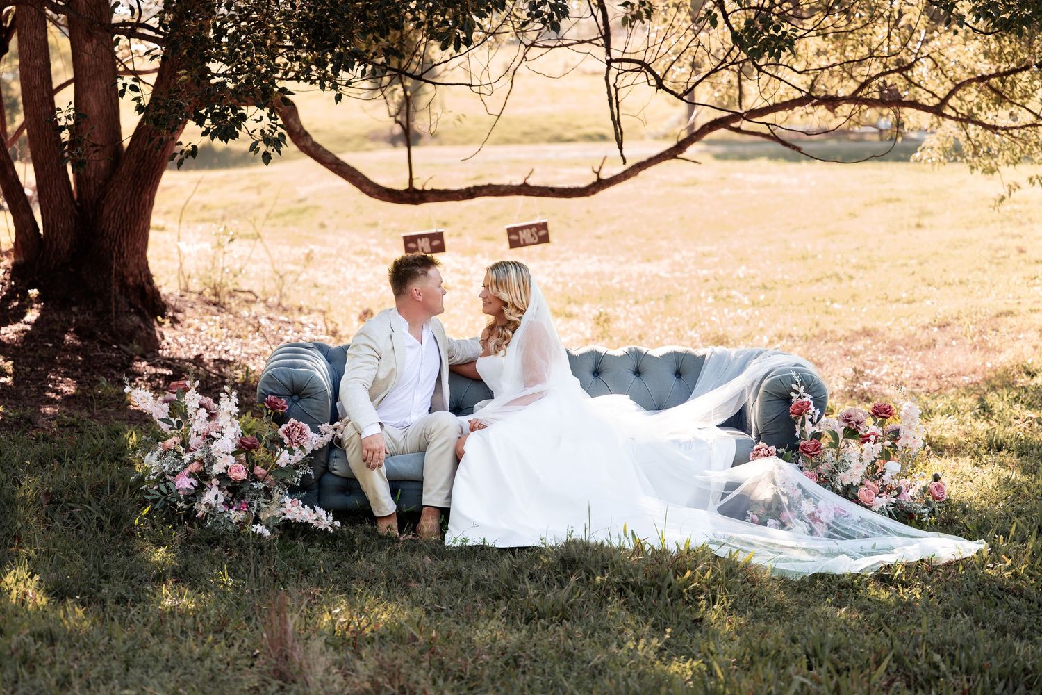 Bride and groom sit on a tufted sofa surrounded by flowers under a tree in a sunlit field.