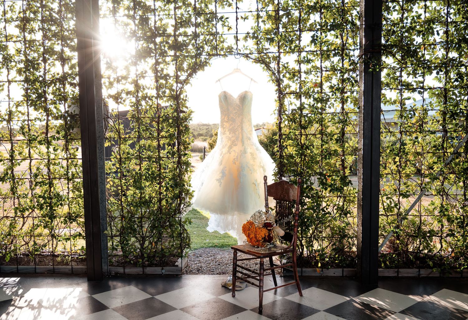 A white wedding dress hangs in front of a sunlit window with greenery visible outside.