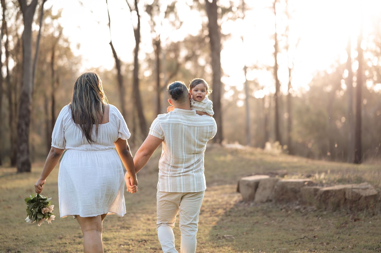 Couple walking hand in hand through a sunlit wooded area wearing coordinated white outfits.