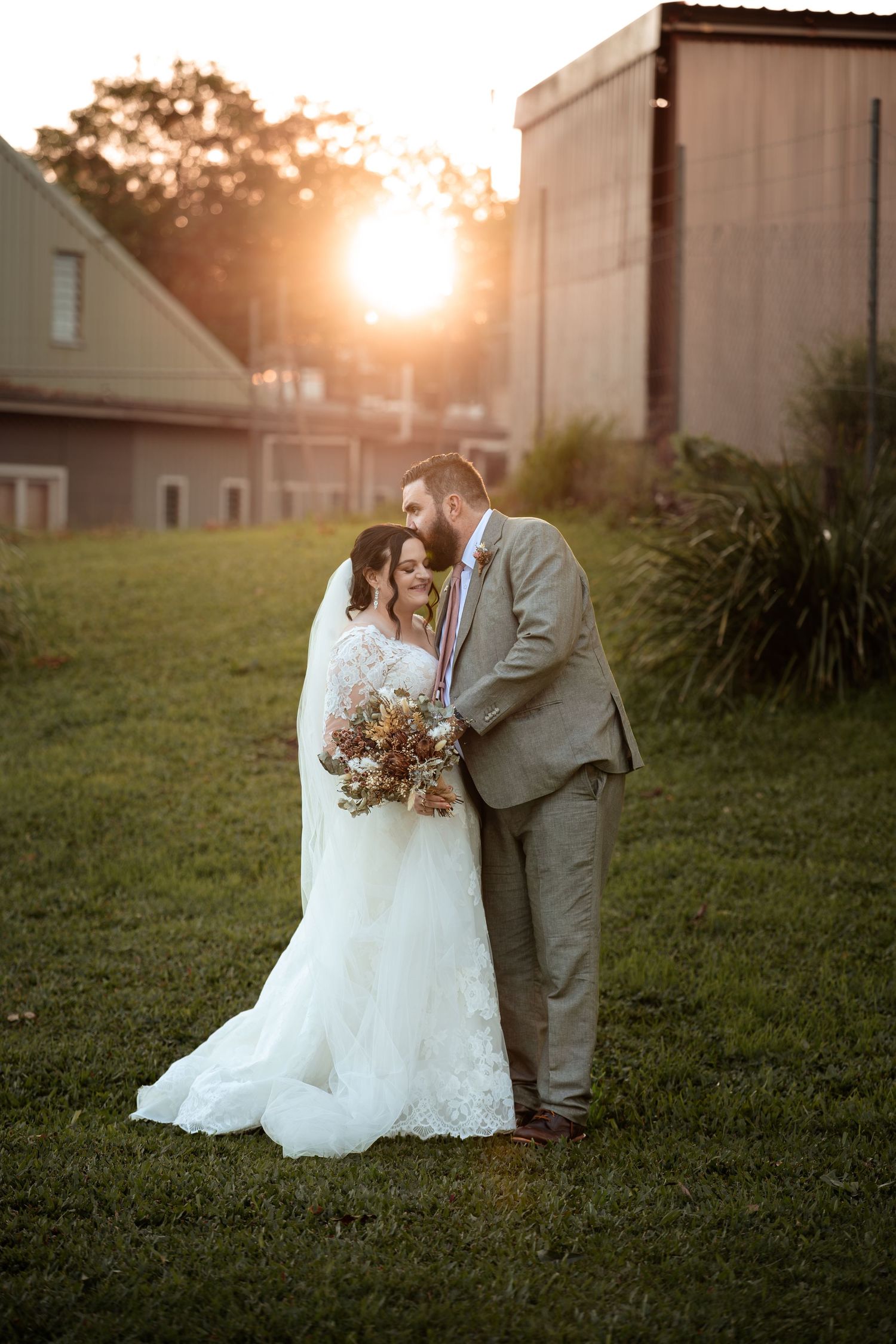 Bride and groom share a romantic sunset moment outdoors, bride holding a bouquet in a lace gown, groom in a gray suit.