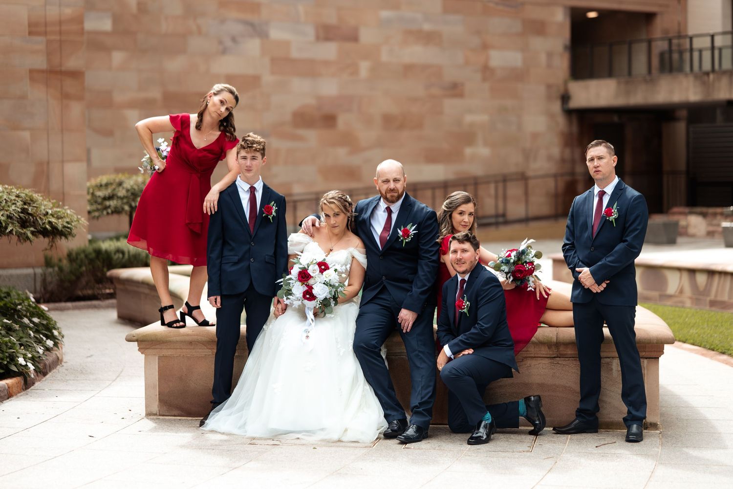 Wedding party in navy suits and red dresses posing playfully outdoors, bride in white gown holding red bouquet.