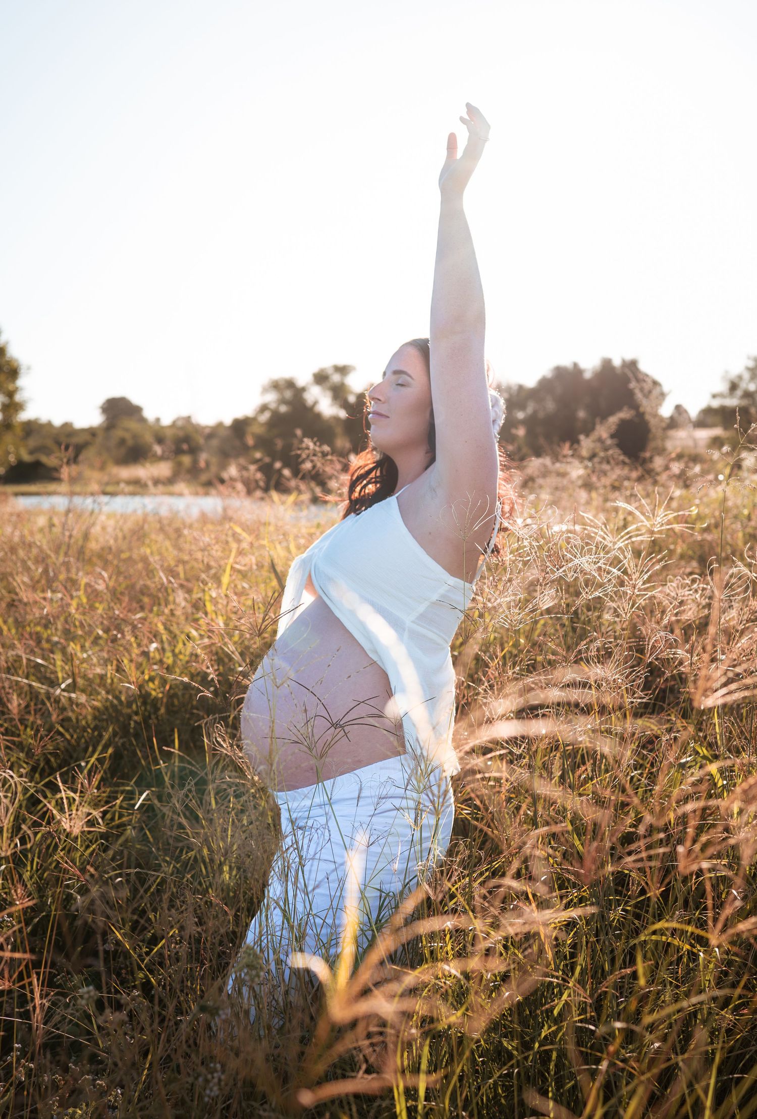 Woman in white dress poses gracefully in golden field during sunset maternity session.