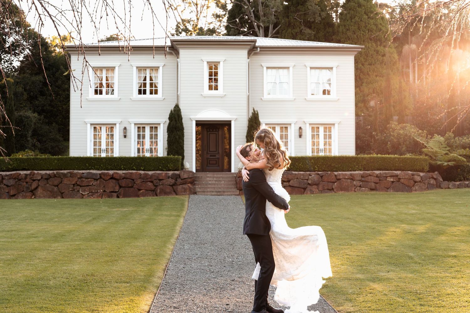 Groom lifting bride in wedding attire outside elegant white manor house with manicured gardens at golden hour.