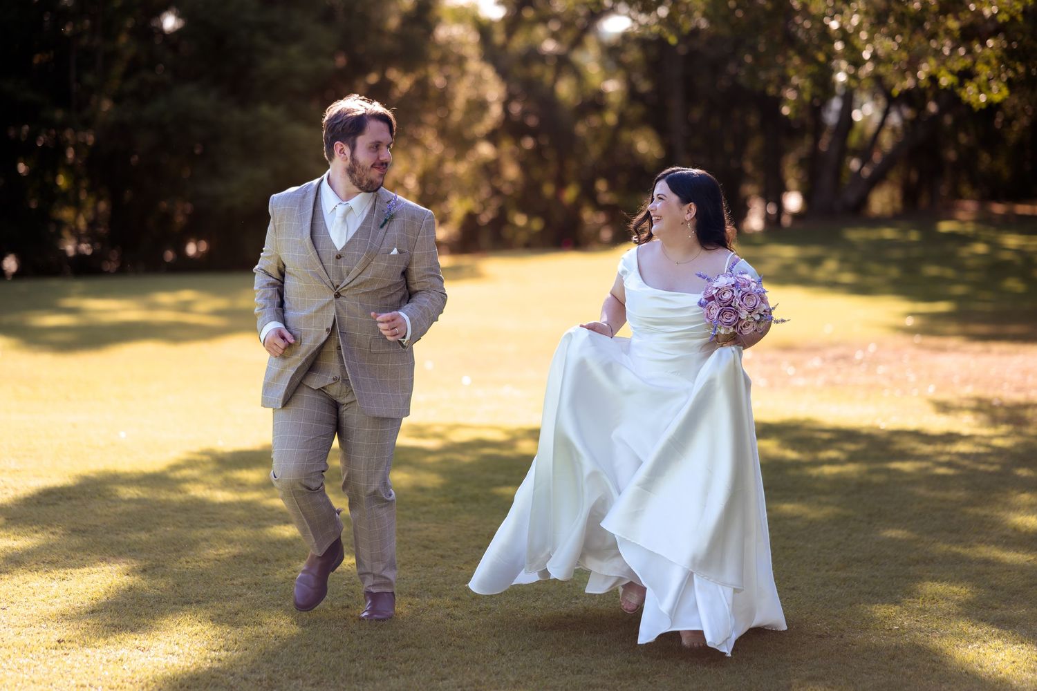 Joyful bride and groom running on green lawn, bride in white gown holding bouquet, groom in tan suit, outdoor wedding photo.