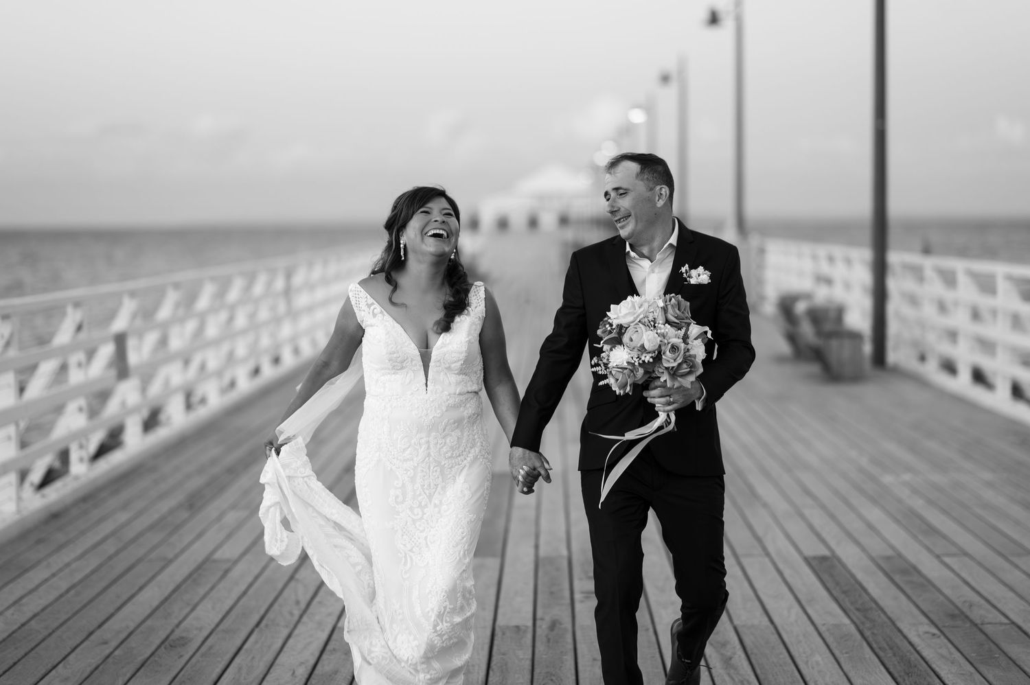 Black and white photo of a bride and groom walking hand in hand on a pier, laughing joyfully on their wedding day.