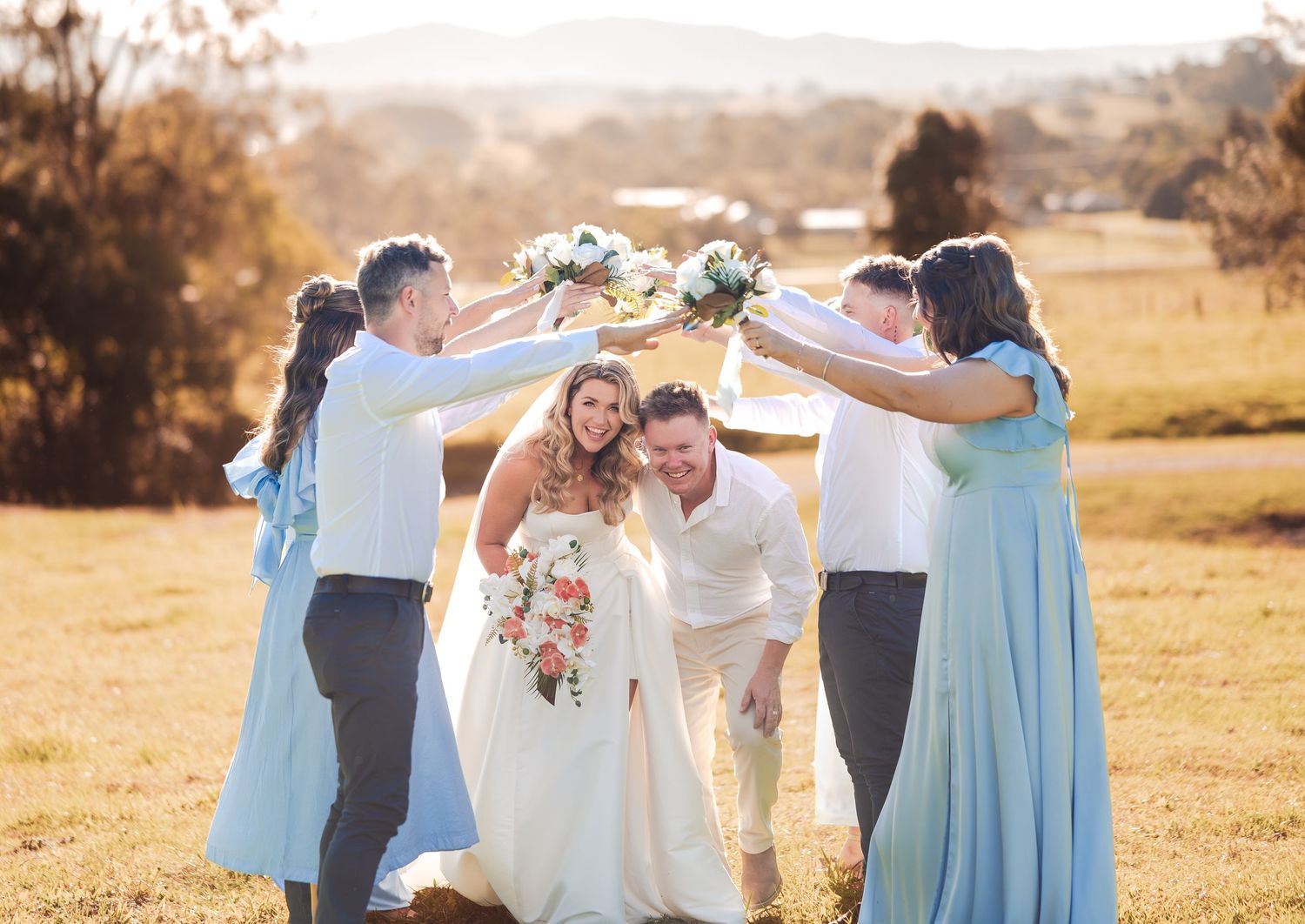 Bride and groom celebrate with bridesmaids in light blue dresses, raising bouquets in a sunny field.