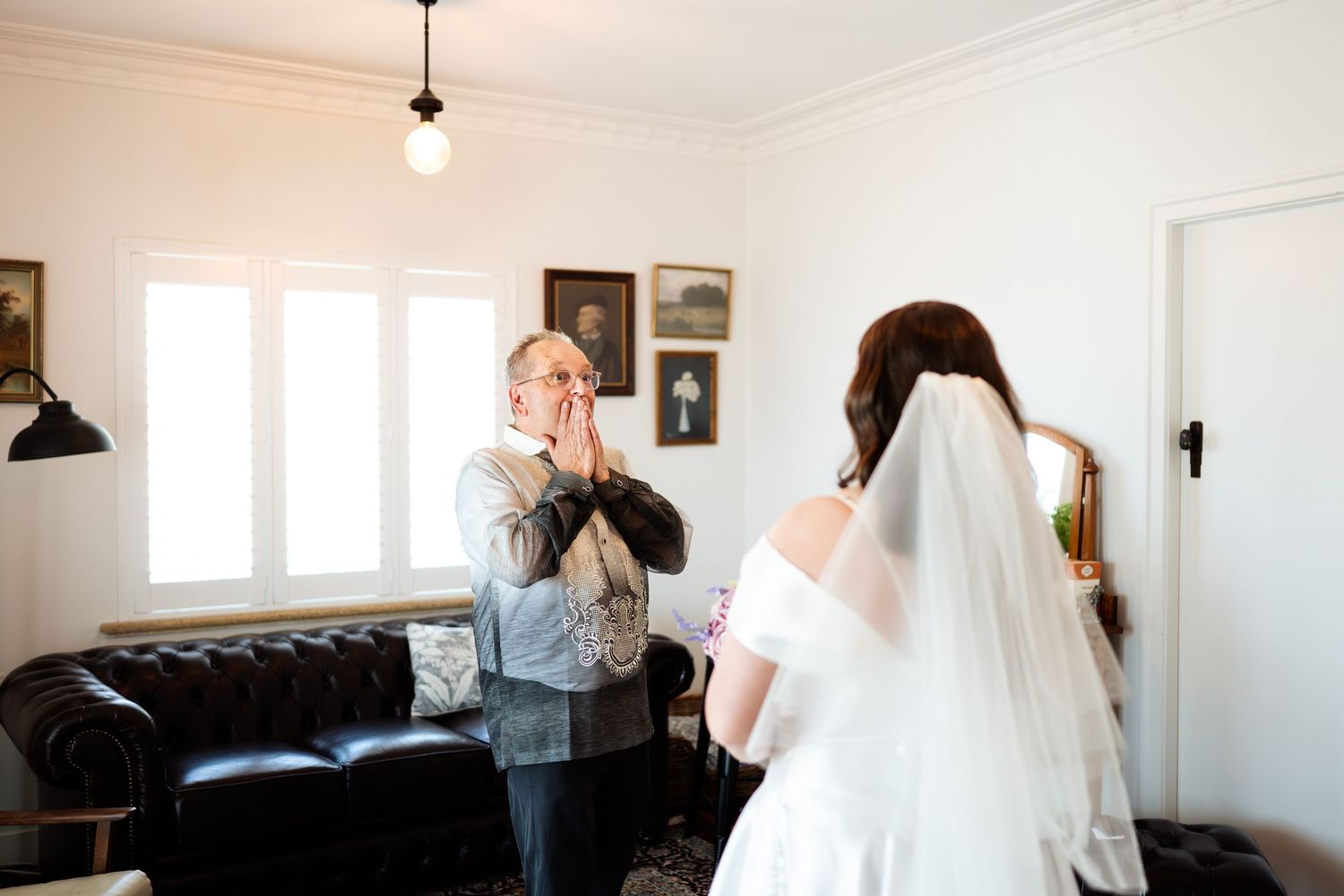 Emotional father clasps hands to face seeing bride daughter in white wedding gown and veil for the first time.