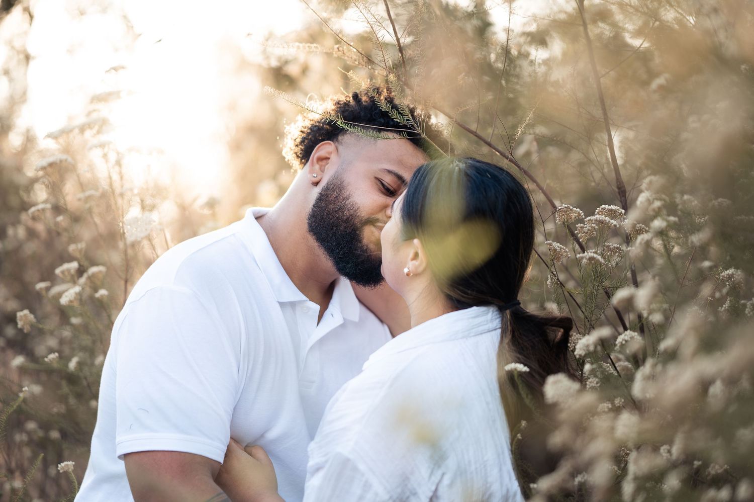 Couple in white clothing embracing in golden sunlight against a natural backdrop.