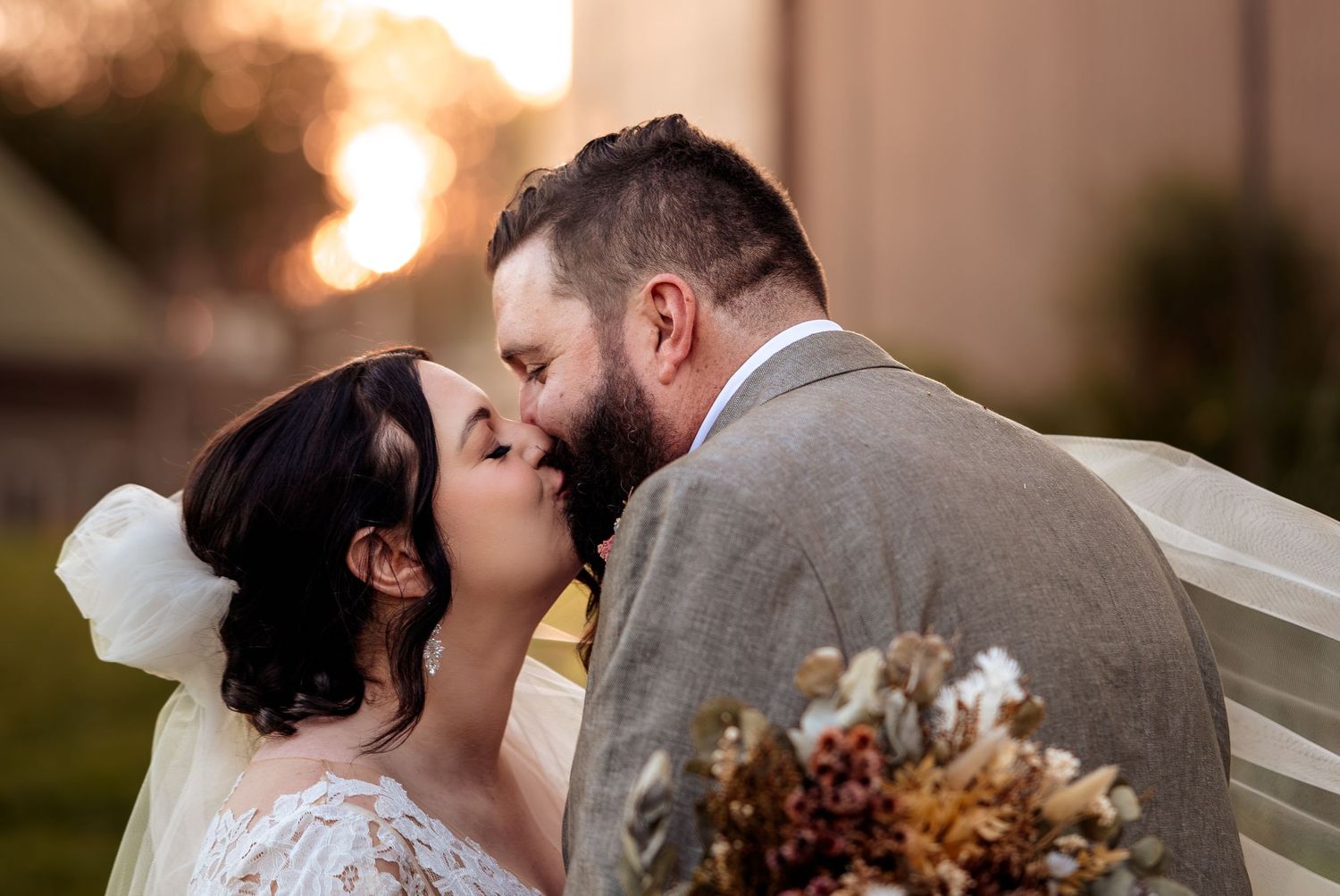 Bride and groom share a romantic kiss at golden hour, with bride holding a rustic dried flower bouquet.