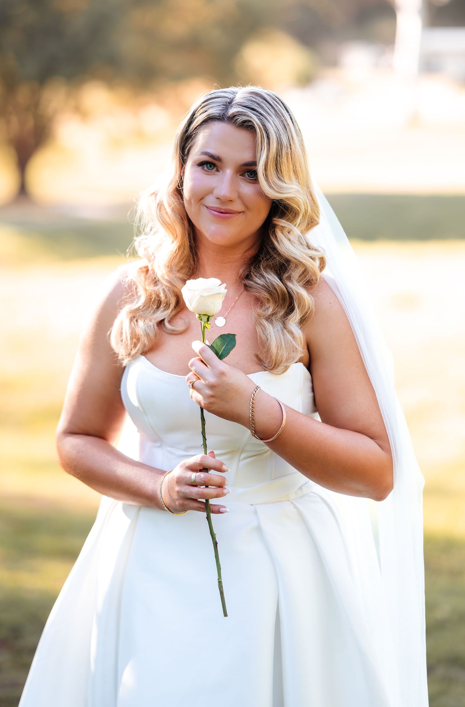 Bride in white strapless gown holding a single white rose, wearing a veil, smiling outdoors on a sunny day.