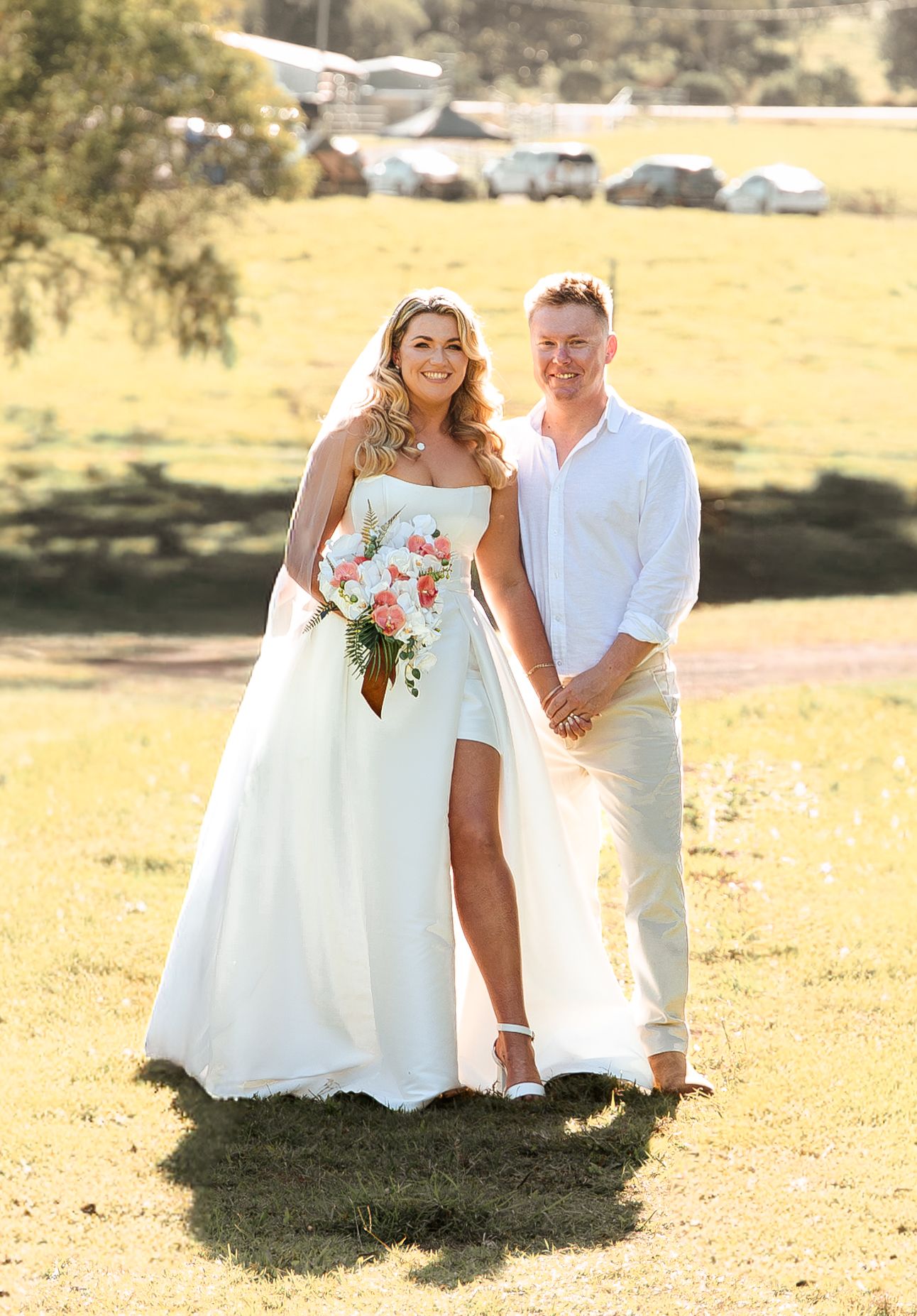 Bride in white gown holding floral bouquet stands with groom in white shirt on green grass at outdoor wedding.