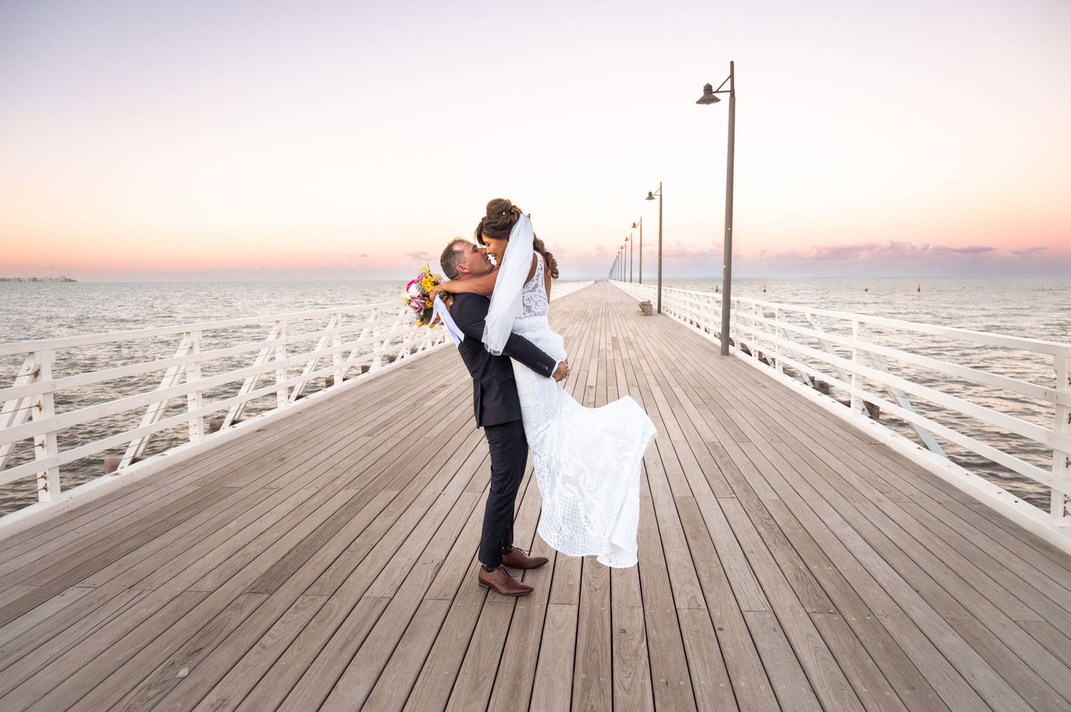 Couple sharing a romantic kiss on a wooden pier at sunset, groom lifting bride in wedding attire.