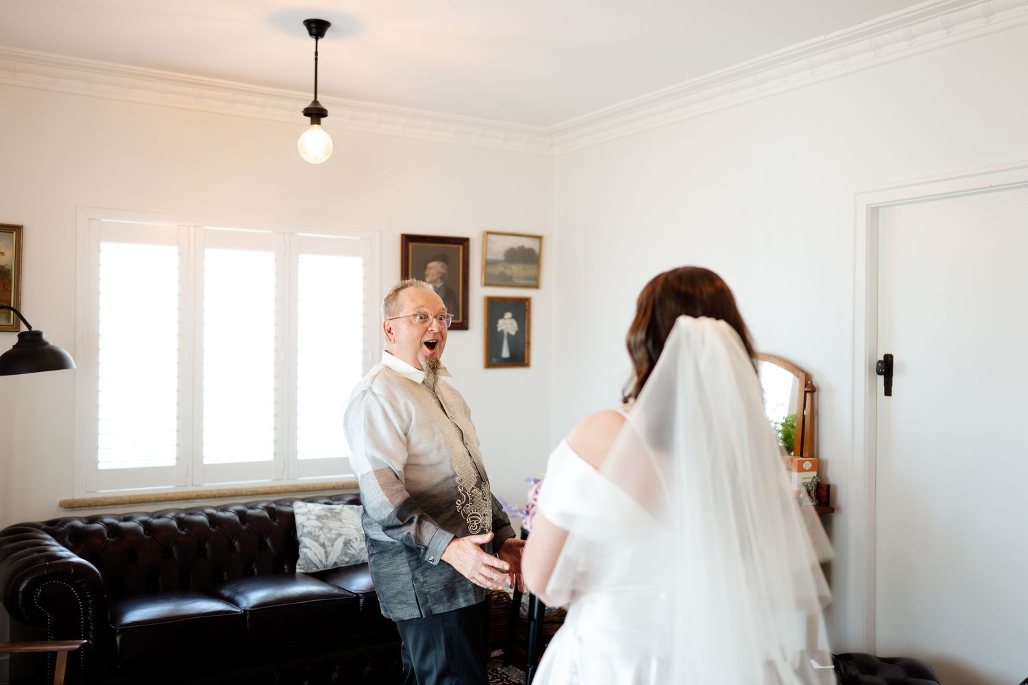 Father reacts with joy seeing bride in wedding dress and veil during getting ready moment indoors.