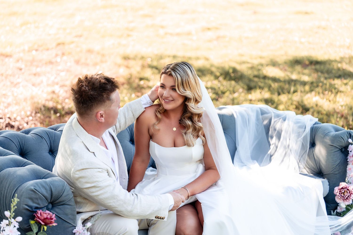 Bride and groom sit together on a blue velvet couch in a sunny field, sharing a tender moment.