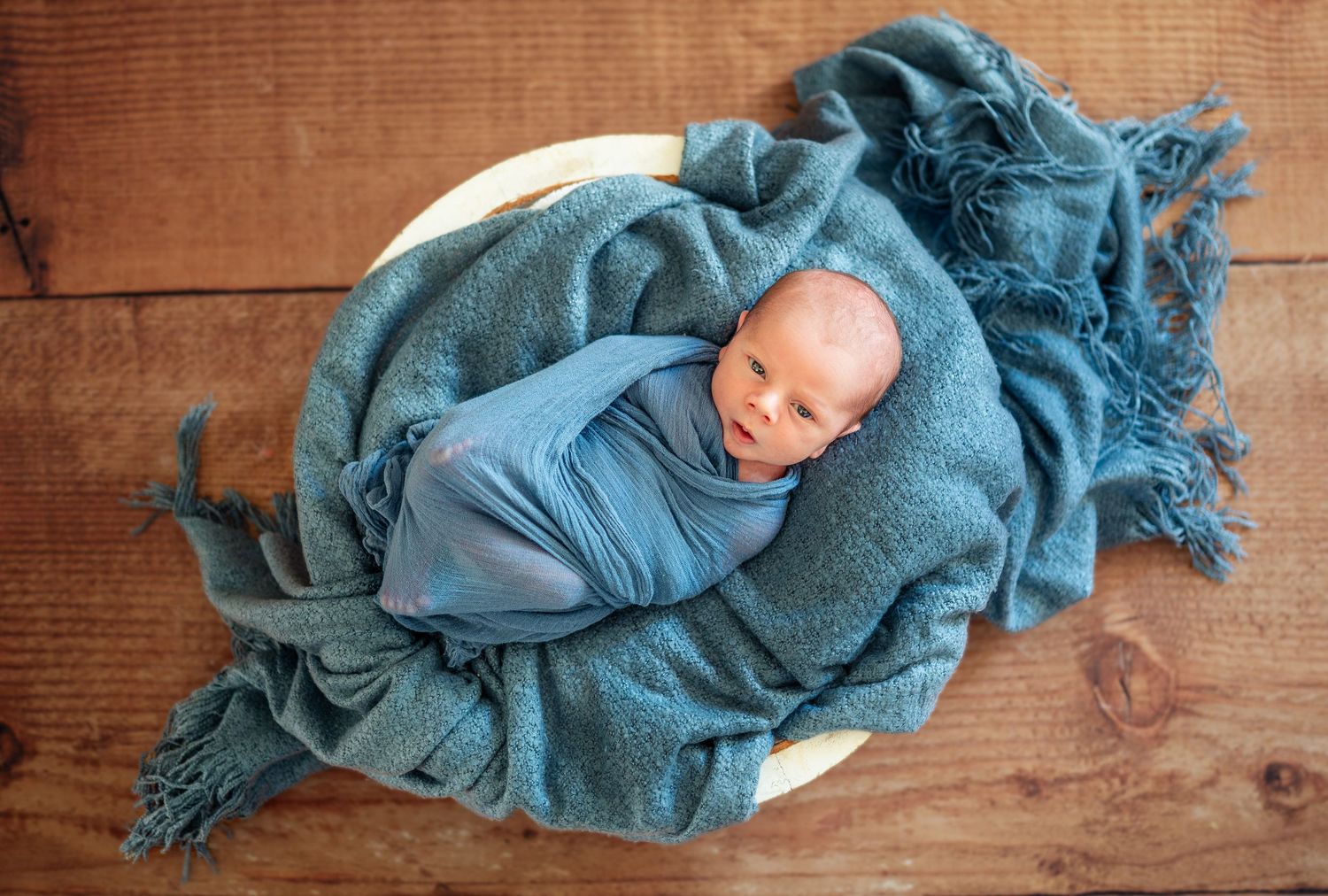 Newborn baby wrapped in teal blanket lying on wooden surface.