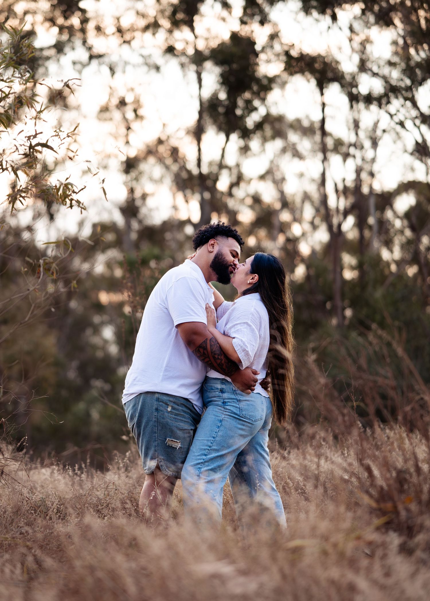 Romantic couple in casual attire sharing intimate moments in tall grass during sunset.