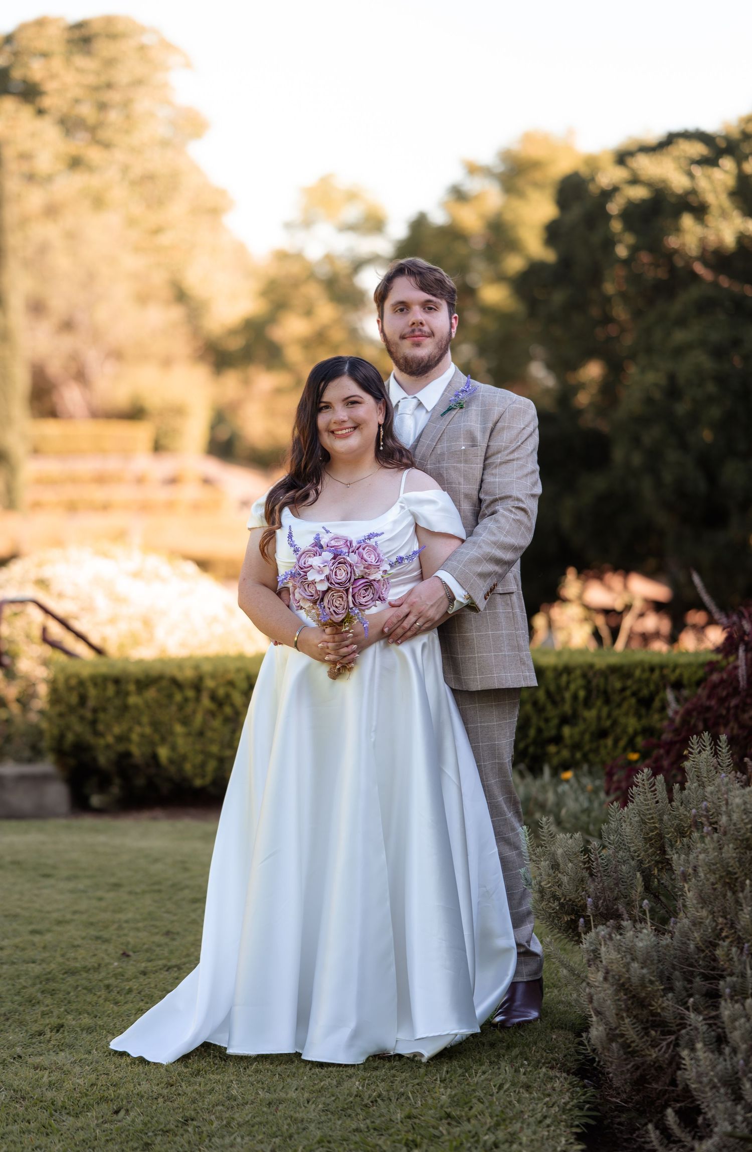 Bride in white gown holding purple bouquet stands with groom in gray suit outdoors in garden setting.