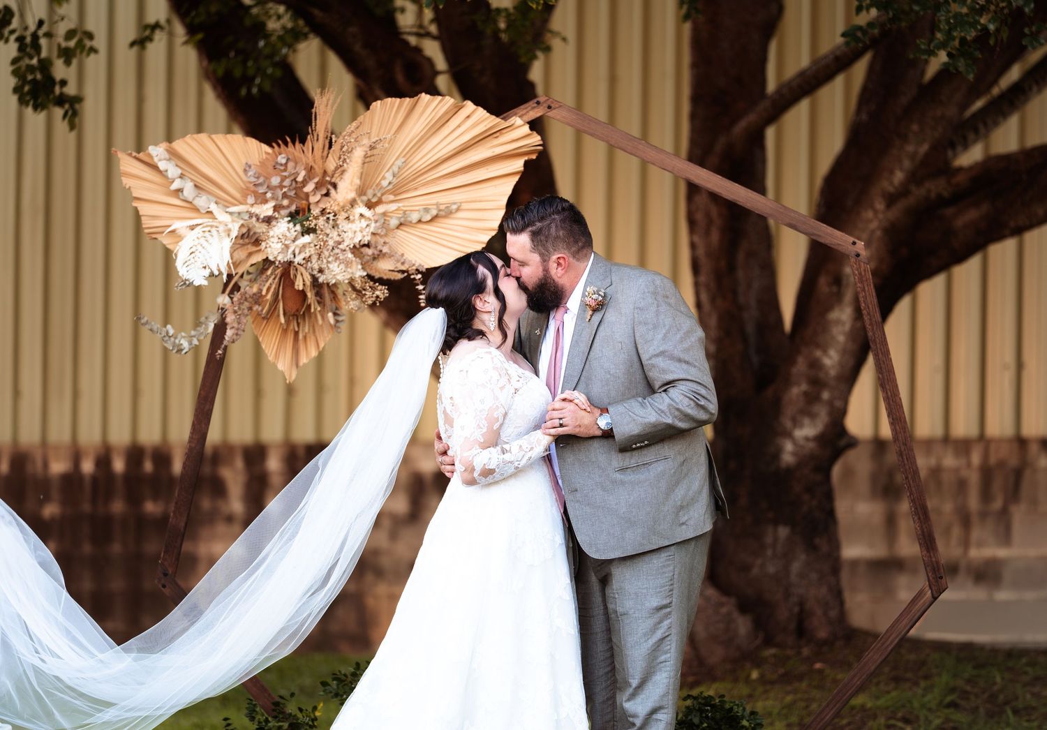 Bride and groom share a kiss under a boho floral arch with dried palm fans at an outdoor wedding ceremony.