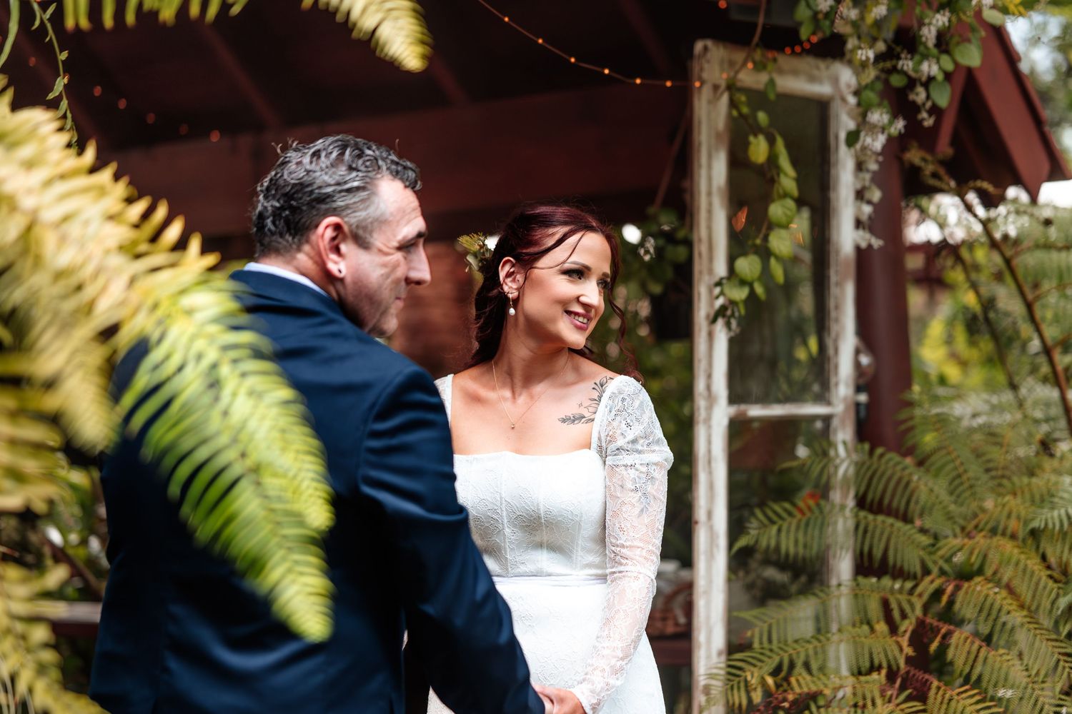 Bride and groom share a tender moment surrounded by lush ferns at an outdoor rustic wedding venue.