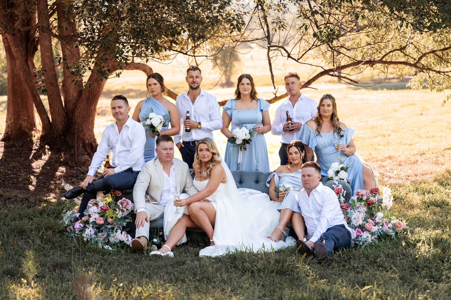 Wedding party posing under tree with colorful flowers, bridesmaids in blue dresses.