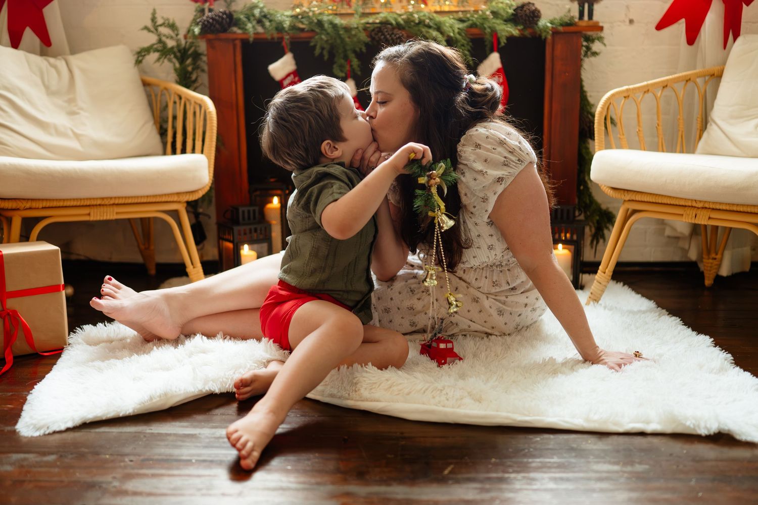 Mother and child sharing a kiss on a white rug by a Christmas fireplace with festive holiday decorations.