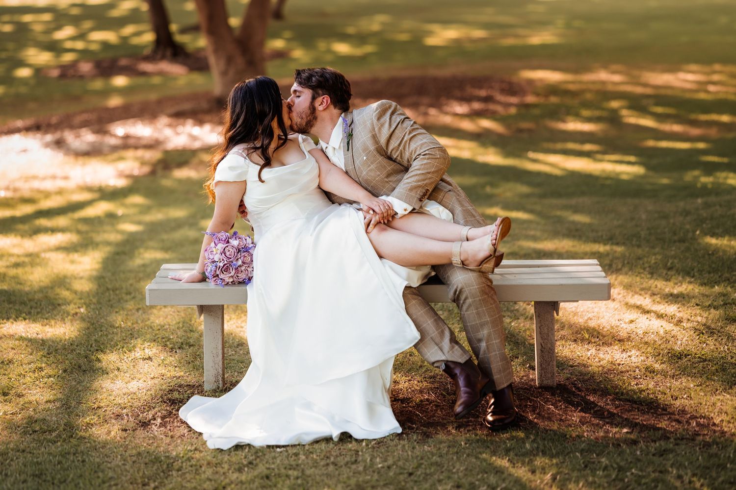 Bride and groom sharing a romantic kiss while seated on a wooden bench in a sunny park.