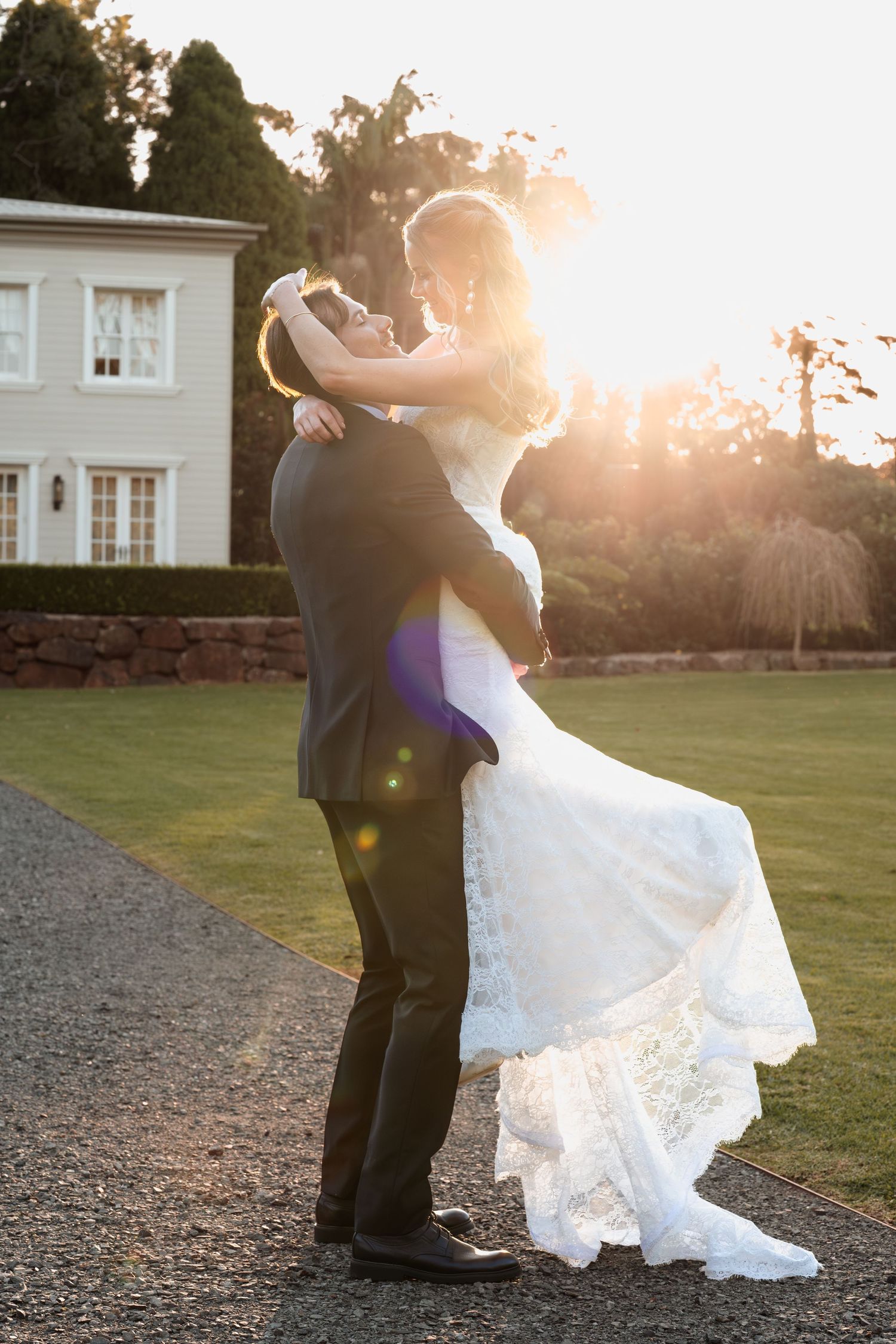 Bride and groom embrace during golden hour sunset at elegant estate venue with white wedding dress flowing.