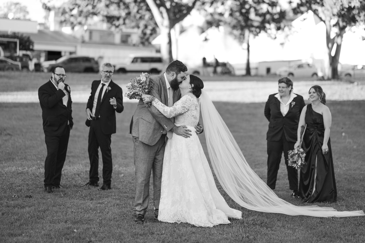 Black and white wedding photo of bride and groom kissing surrounded by bridal party on green lawn.