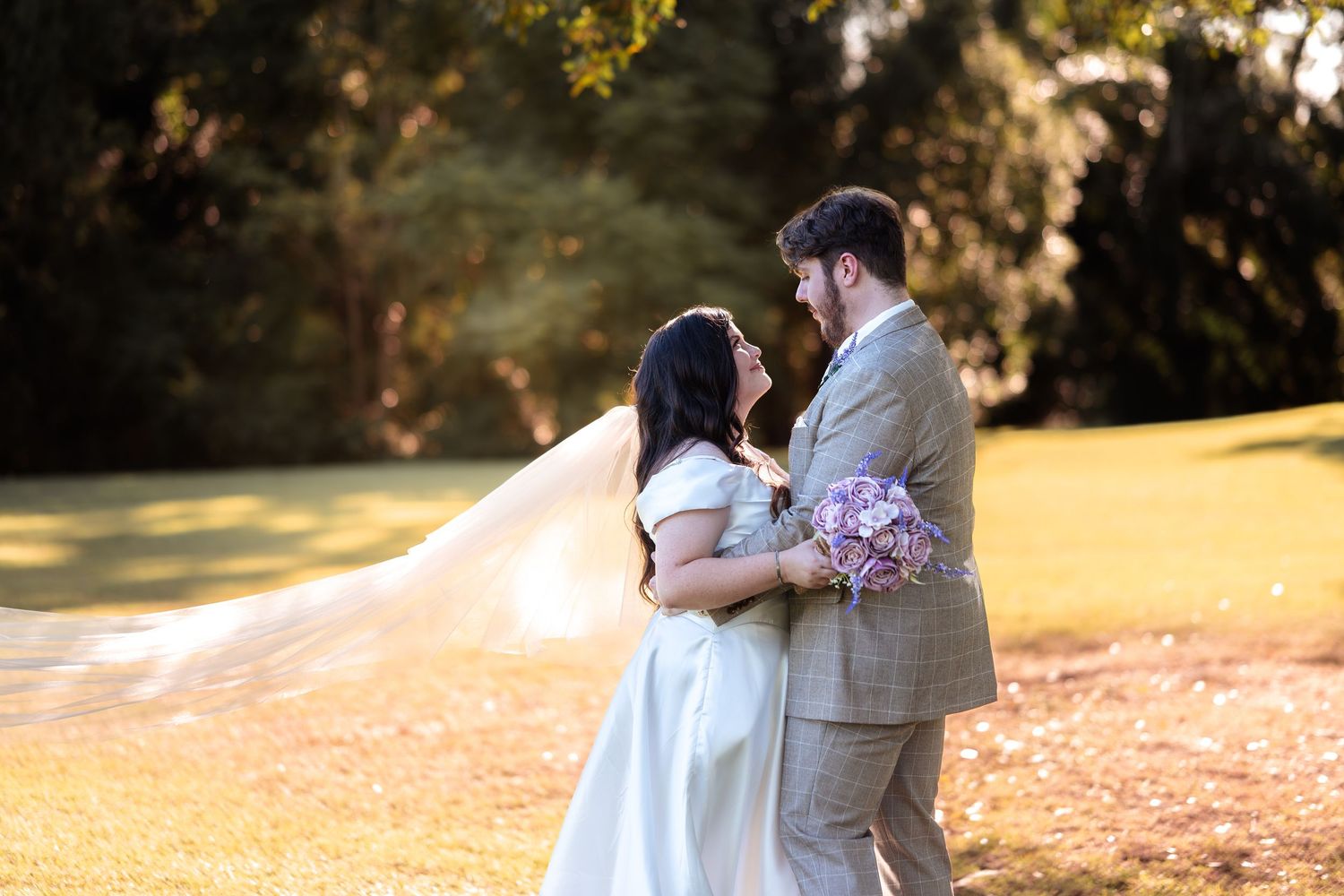 Bride and groom share a romantic moment outdoors, her veil flowing in the wind as she holds a purple bouquet.