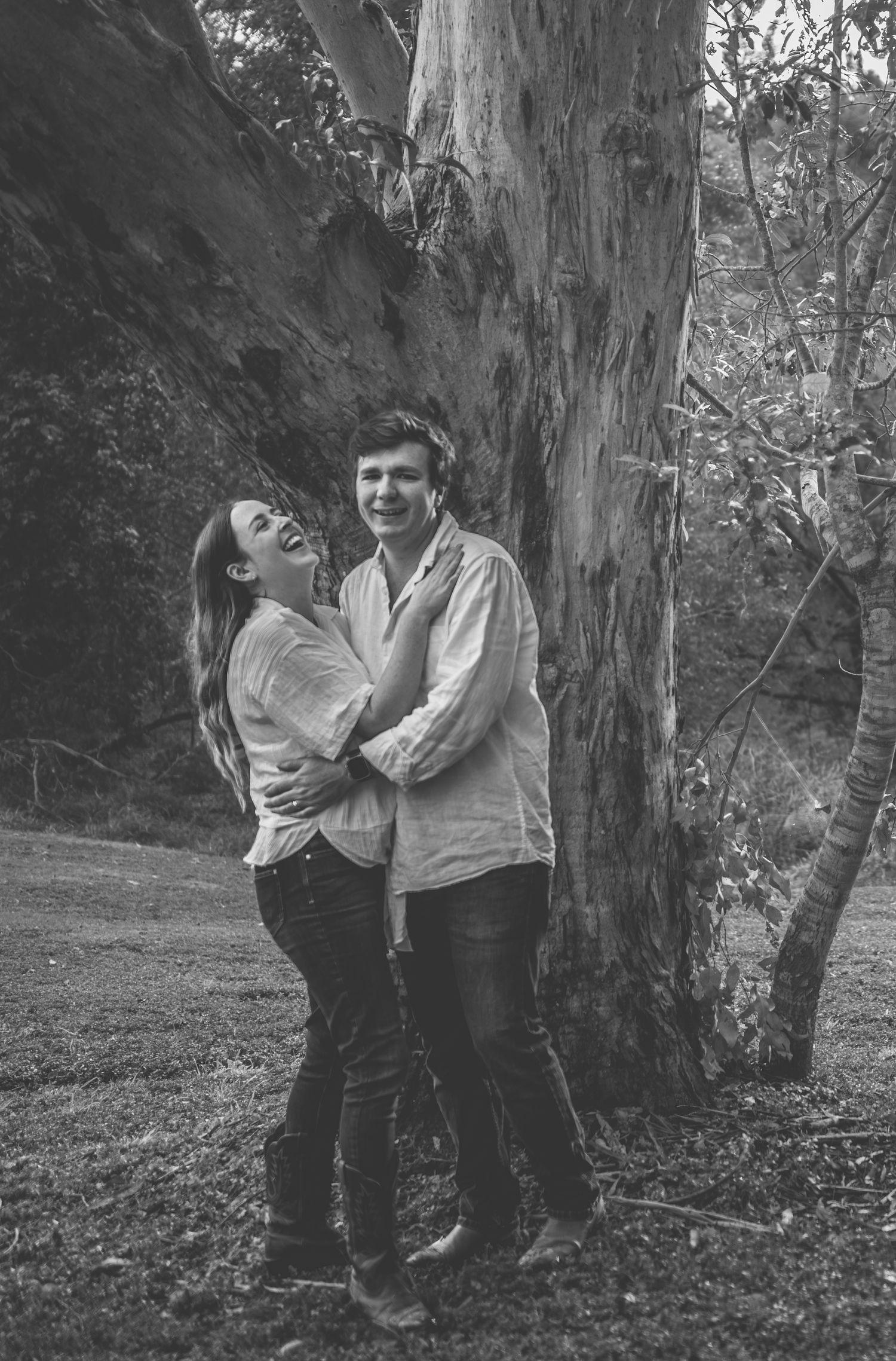 Black and white photo of couple embracing by large tree trunk.
