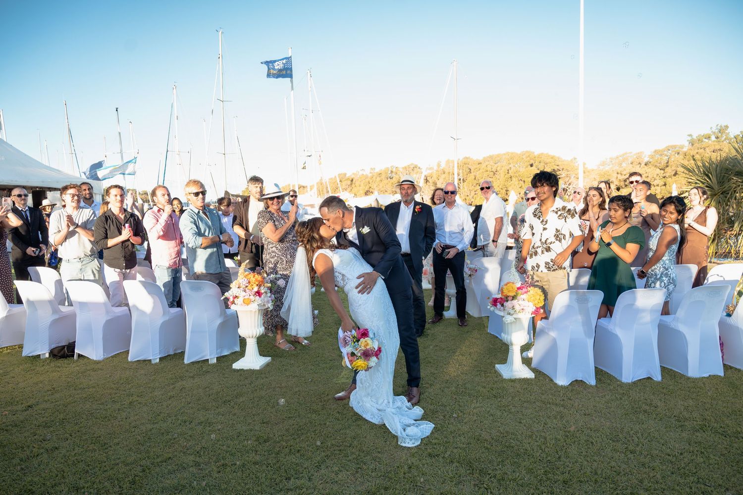 Bride and groom share a romantic dip kiss at outdoor wedding ceremony by marina with guests applauding.