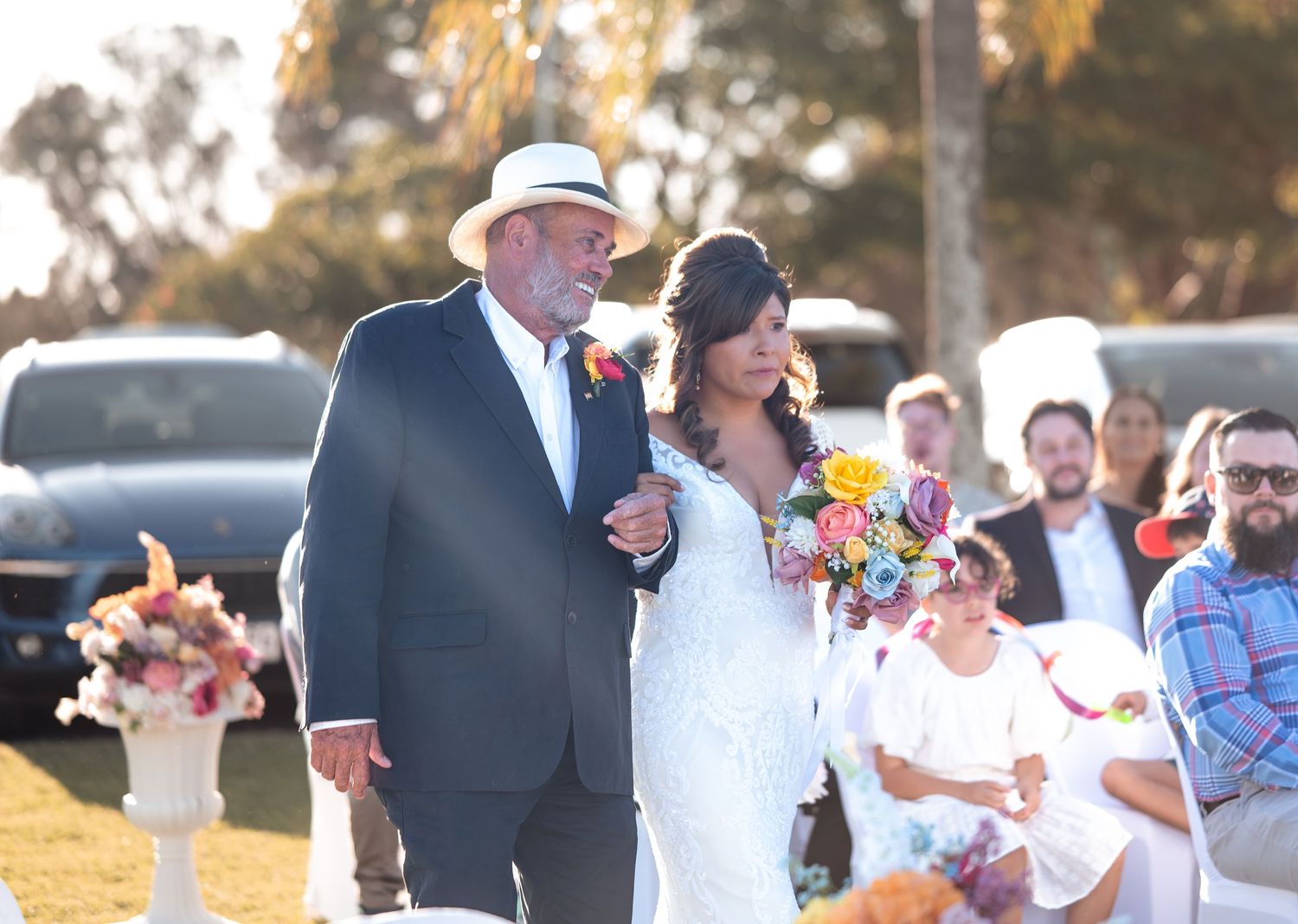 Bride in white lace gown walks down outdoor aisle with older man in suit and hat at sunny wedding ceremony.