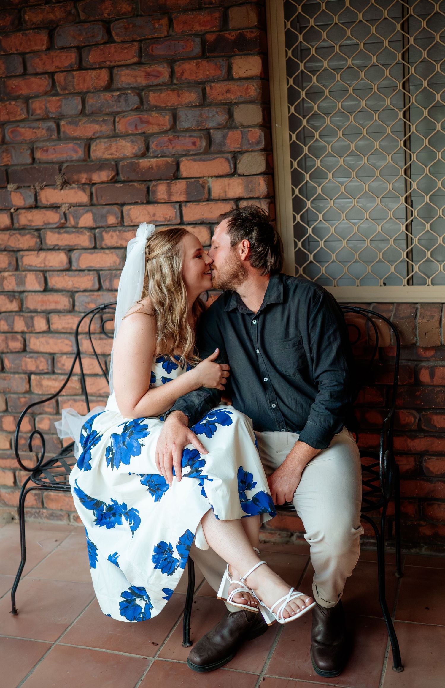 A romantic couple sharing a kiss while seated on a bench against a rustic brick wall.