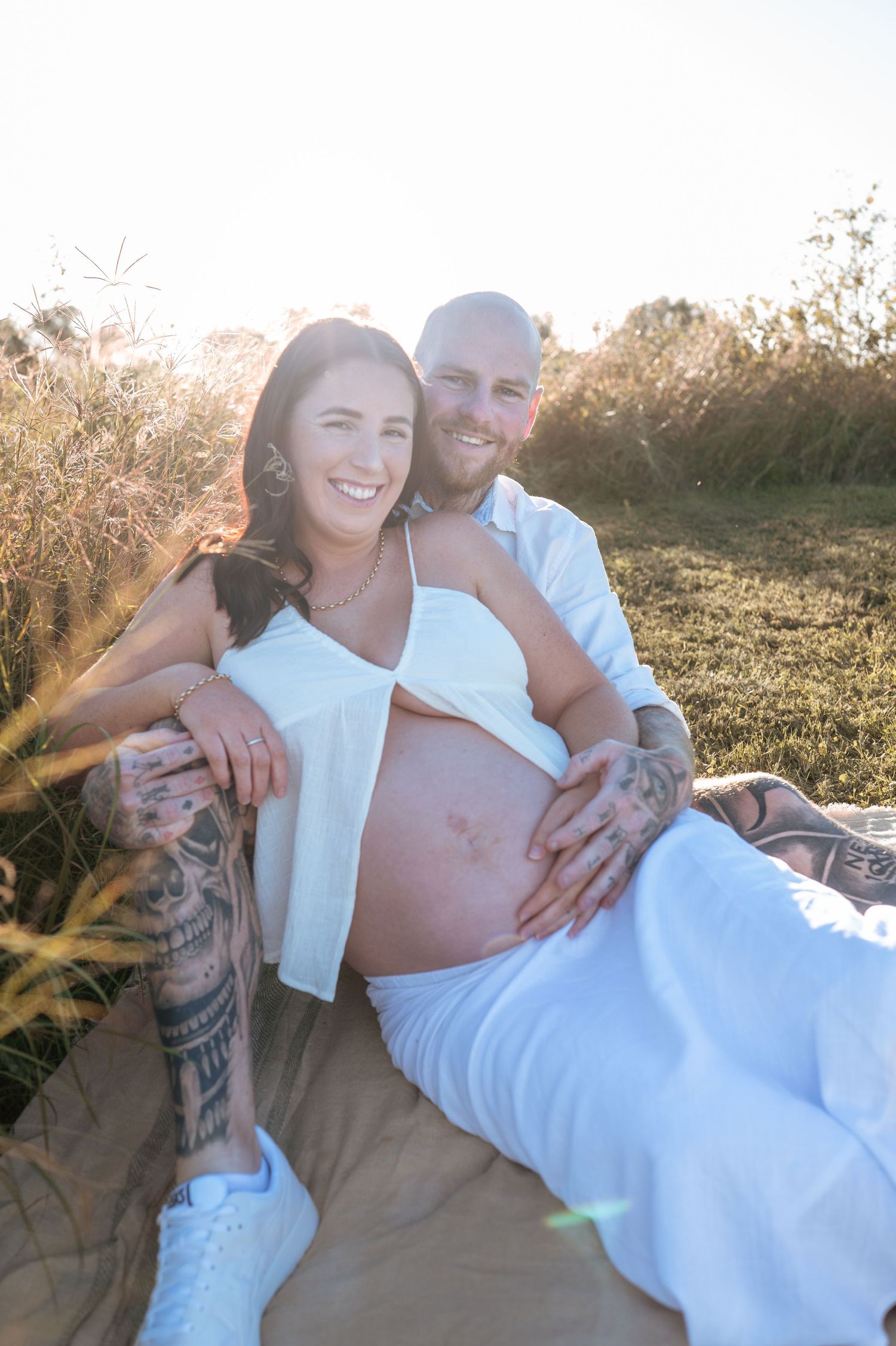 Intimate maternity photoshoot of couple in white clothing laying on grass during golden hour.