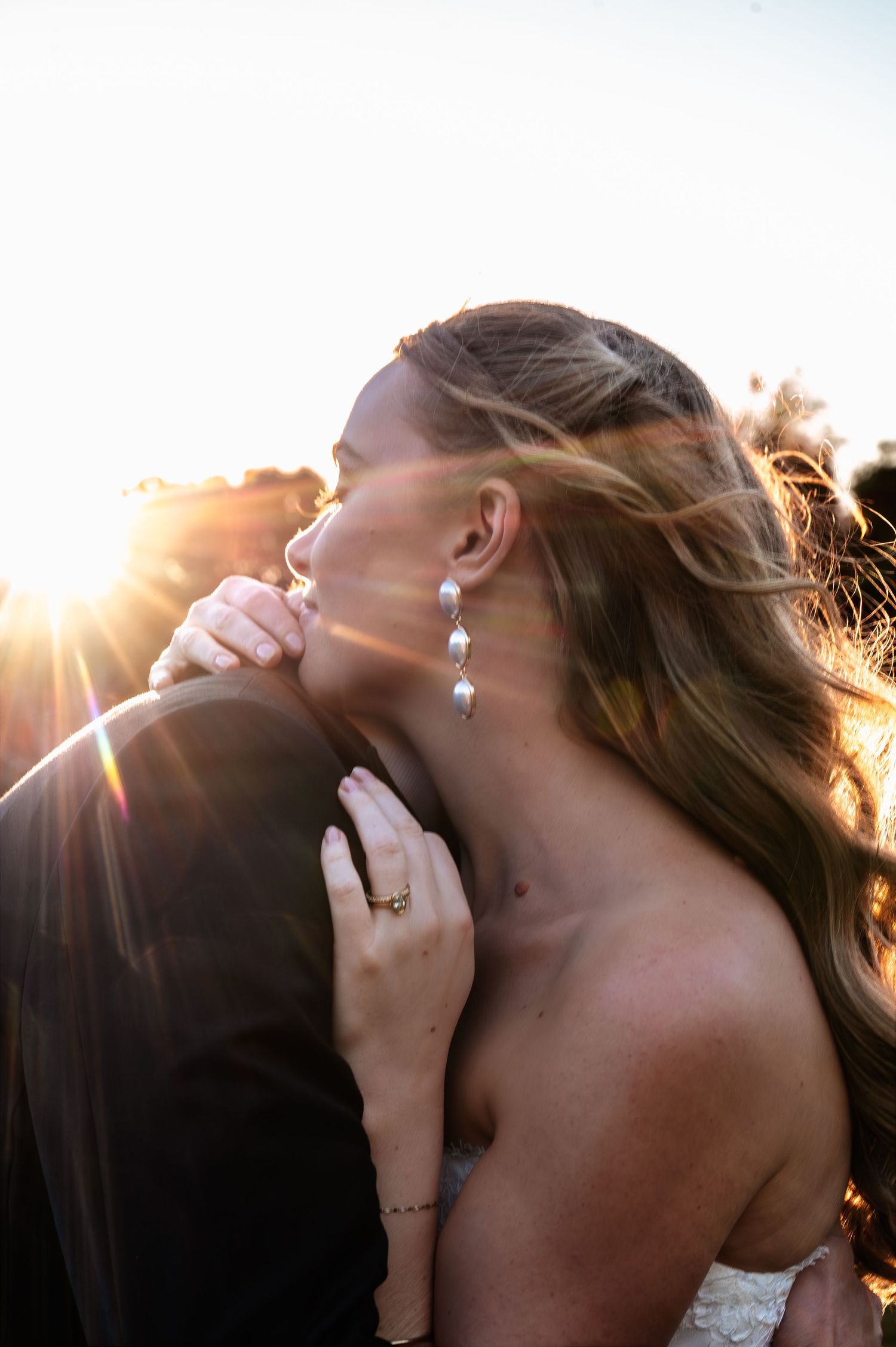Bride and groom kissing at sunset with golden sunlight, bride wearing white dress and earrings with visible engagement ring.