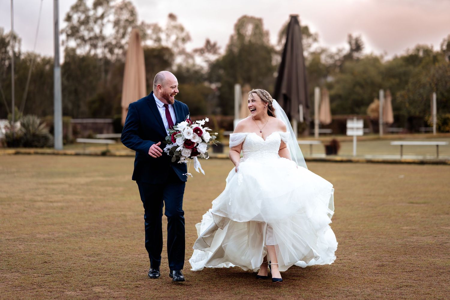 Happy bride and groom walking together outdoors, bride lifting her white off-shoulder wedding gown.